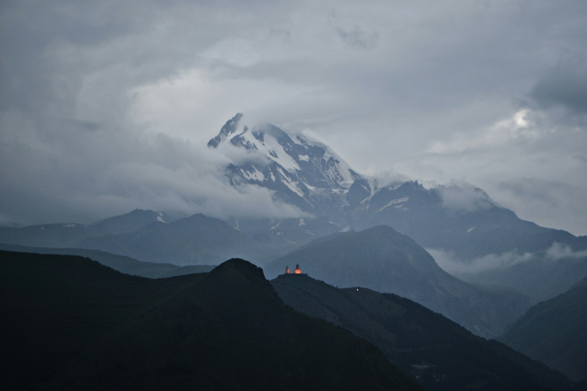 Gergeti Trinity Church lit on its ridge at dusk beneath the clouded bulk of Mount Kazbek