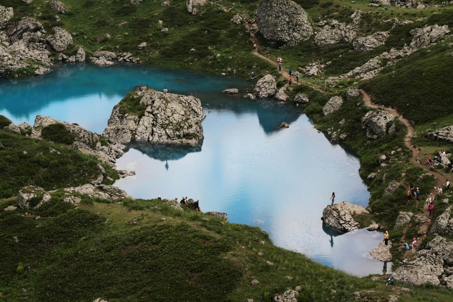 Hikers on the rocky shore and trail around the turquoise Blue Lake at Abudelauri, a boulder island in the middle reflecting the sky