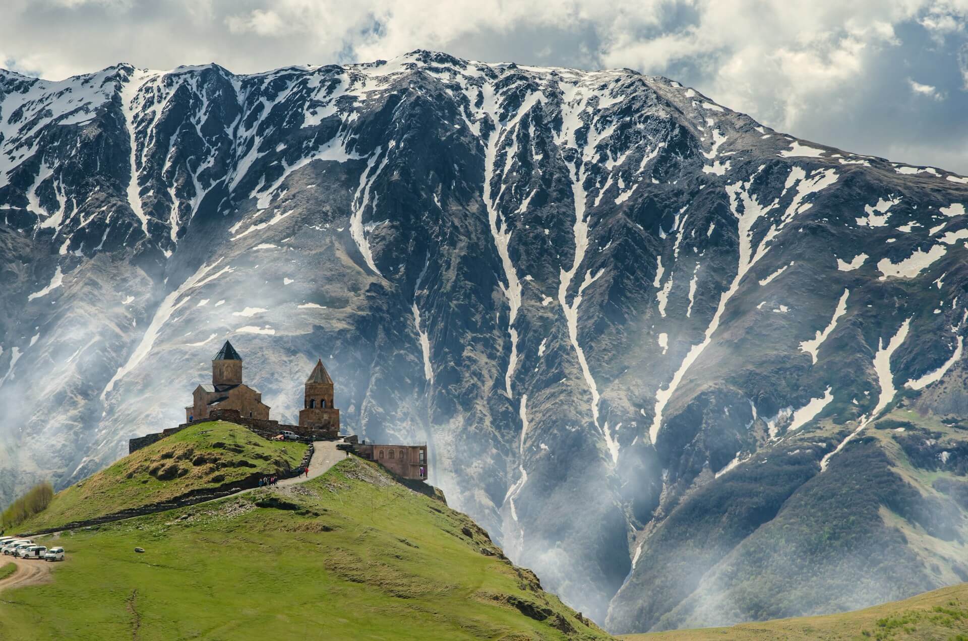 Gergeti Trinity Church on its green hilltop in daylight, in front of a snow-streaked Caucasus ridge with mist rising from the valley
